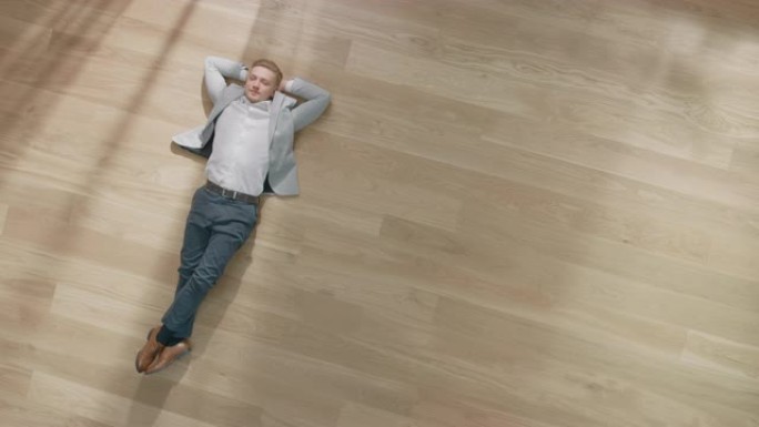 年轻人躺在公寓的木地板上。他穿着夹克和白衬衫。舒适的客厅配有现代简约的内部和木质镶木地板。顶视图摄像