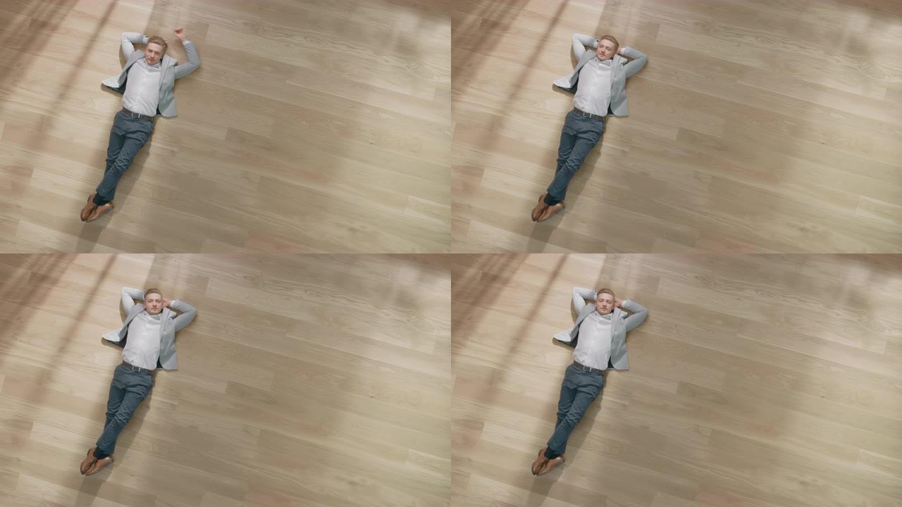 年轻人躺在公寓的木地板上。他穿着夹克和白衬衫。舒适的客厅配有现代简约的内部和木质镶木地板。顶视图摄像