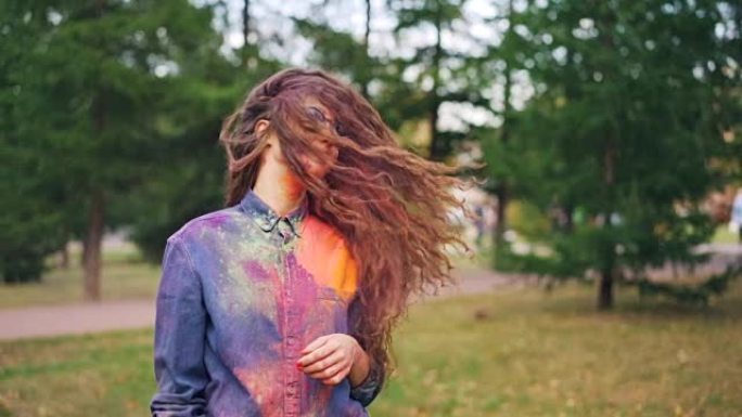 可爱的年轻女子学生的慢动作肖像在洒红节上玩得很开心，独自一人站在公园外面，把她的头发涂上五颜六色的粉