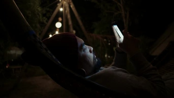 时髦的男人晚上在吊床上用智能手机放松。