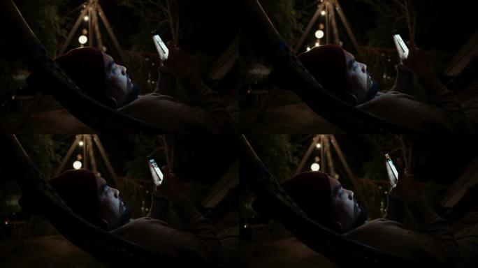 时髦的男人晚上在吊床上用智能手机放松。