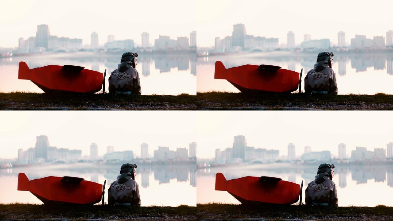 小飞行员男孩坐在令人惊叹的宁静城市湖全景附近，带着有趣的红色纸板飞机慢动作。