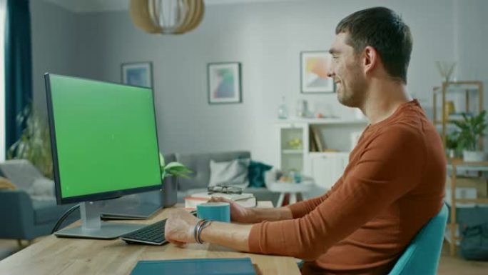 英俊的微笑男人坐在家里的办公桌旁，使用带有绿屏模型的个人计算机。他住在舒适的公寓里，室内有现代客厅。