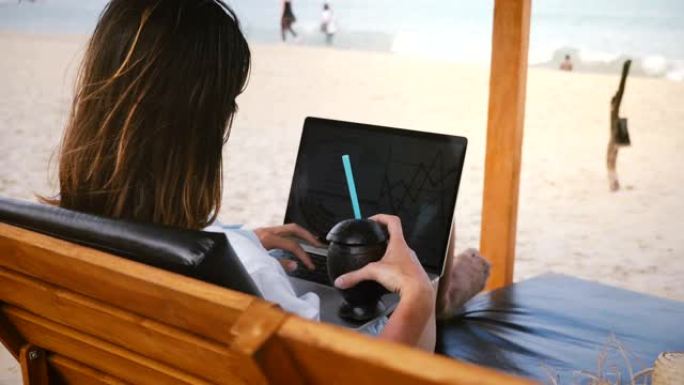 在风景秀丽的海洋海滩的躺椅上，成功的快乐自由职业者在笔记本电脑图表上喝酒。