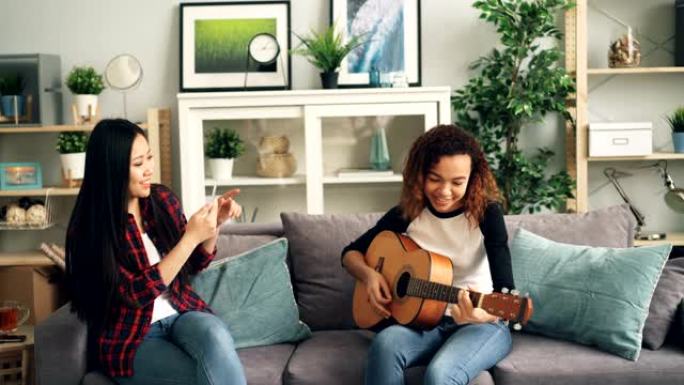 女性朋友在家里玩得开心，非洲裔美国女孩在弹吉他，亚洲年轻女子在用智能手机录制视频。友谊和音乐概念。