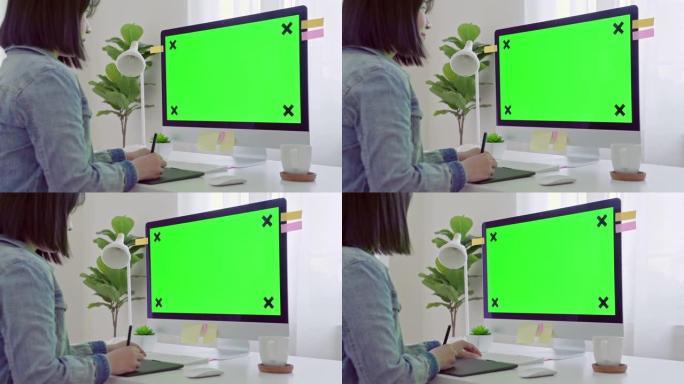 年轻的成年人在办公桌前的计算机上工作，该计算机带有模拟绿屏。在肩膀上的镜头。她坐在家里的桌子旁。