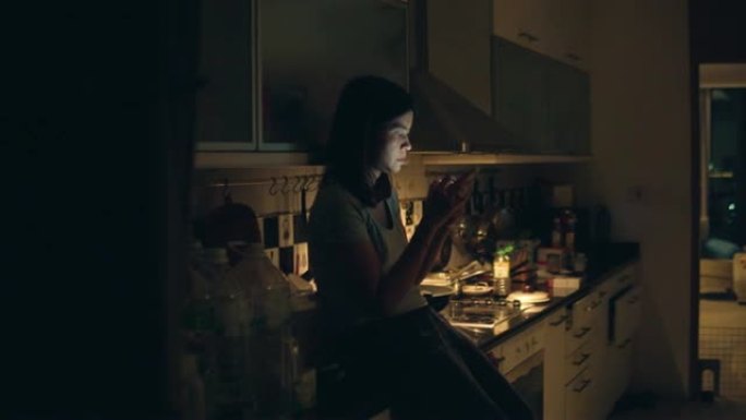 年轻女子坐在厨房柜台上，使用智能手机站在家里的厨房里午夜。
