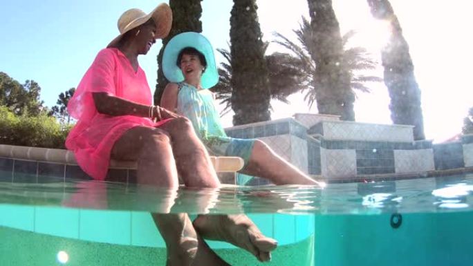 两名多种族的高级妇女在游泳池旁，脚在水中