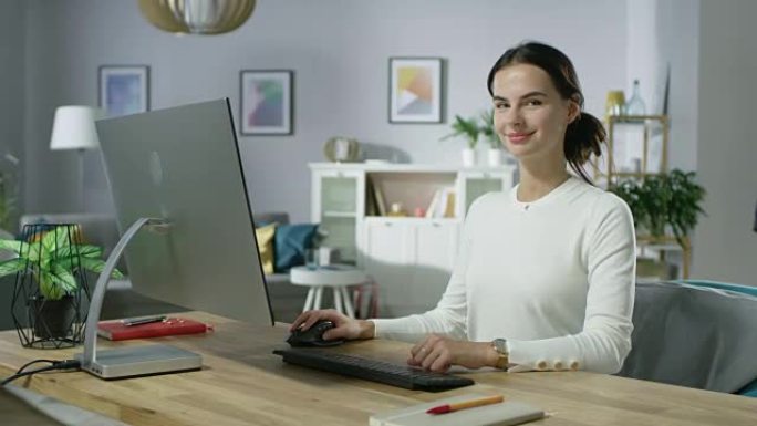 缩小美丽的年轻女子在舒适的客厅在个人电脑上工作的肖像照片。她看着镜头迷人地微笑着。