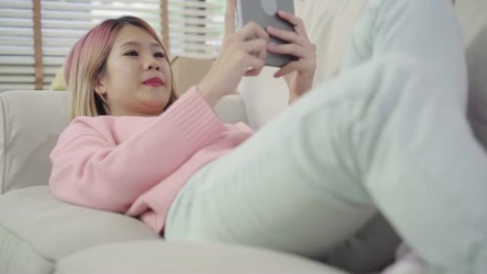 年轻的亚洲女性搬到新家，美丽的女性使用平板电脑社交媒体躺在沙发上放松后，在家里的客厅搬回家。享受时间