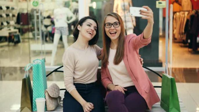 漂亮的年轻女士正在坐在购物中心里休息和娱乐的智能手机上一起自拍。女孩们用手势拥抱和摆姿势。