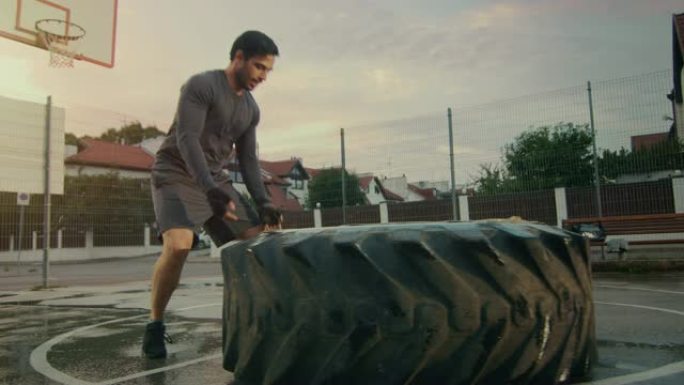 穿着运动服和手套的强壮肌肉健康的年轻人在一个有围栏的室外篮球场上做运动。他在雨后的下午环境中翻转一个
