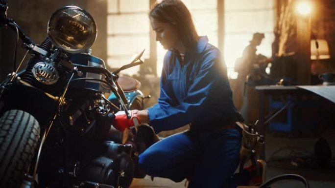 年轻漂亮的女机械师正在设计一辆定制的Bobber摩托车。穿着蓝色连体裤的天才女孩。她用扳手拧紧螺母螺