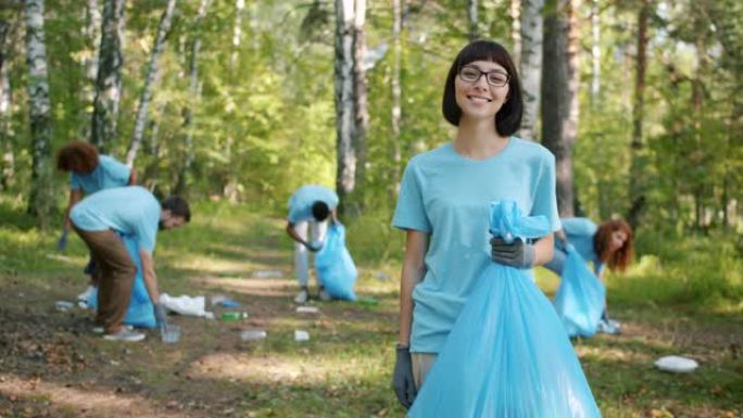 漂亮的女志愿者在污染的公园里看着拿着垃圾袋的相机