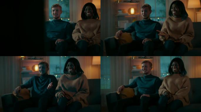 快乐的多元化年轻夫妇坐在沙发上看电视喜剧，他们笑着享受表演。英俊的高加索男孩和黑人女孩在一起。移动摄