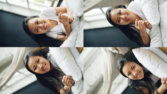 美丽的亚洲黑发女人在床上滚动，带着愉快的微笑看着相机，玩得开心，玩得开心。公寓和千禧一代的概念。