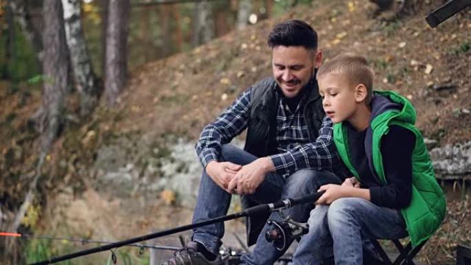 帅哥爱爸爸正在教他的儿子一起坐在河岸上抓鱼，男孩拿着棍子和爸爸说话。爱好、冒险和家庭概念。