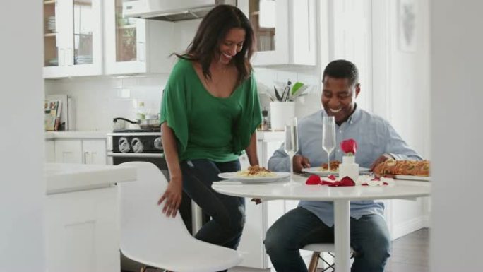 中年黑人坐在厨房里，而他的伴侣为他们提供浪漫的饭菜