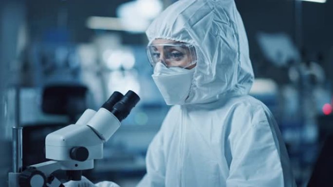 研究工厂洁净室: 工程师/科学家穿着工作服，使用显微镜检查样品，为医疗和高精度电子行业开发高科技现代