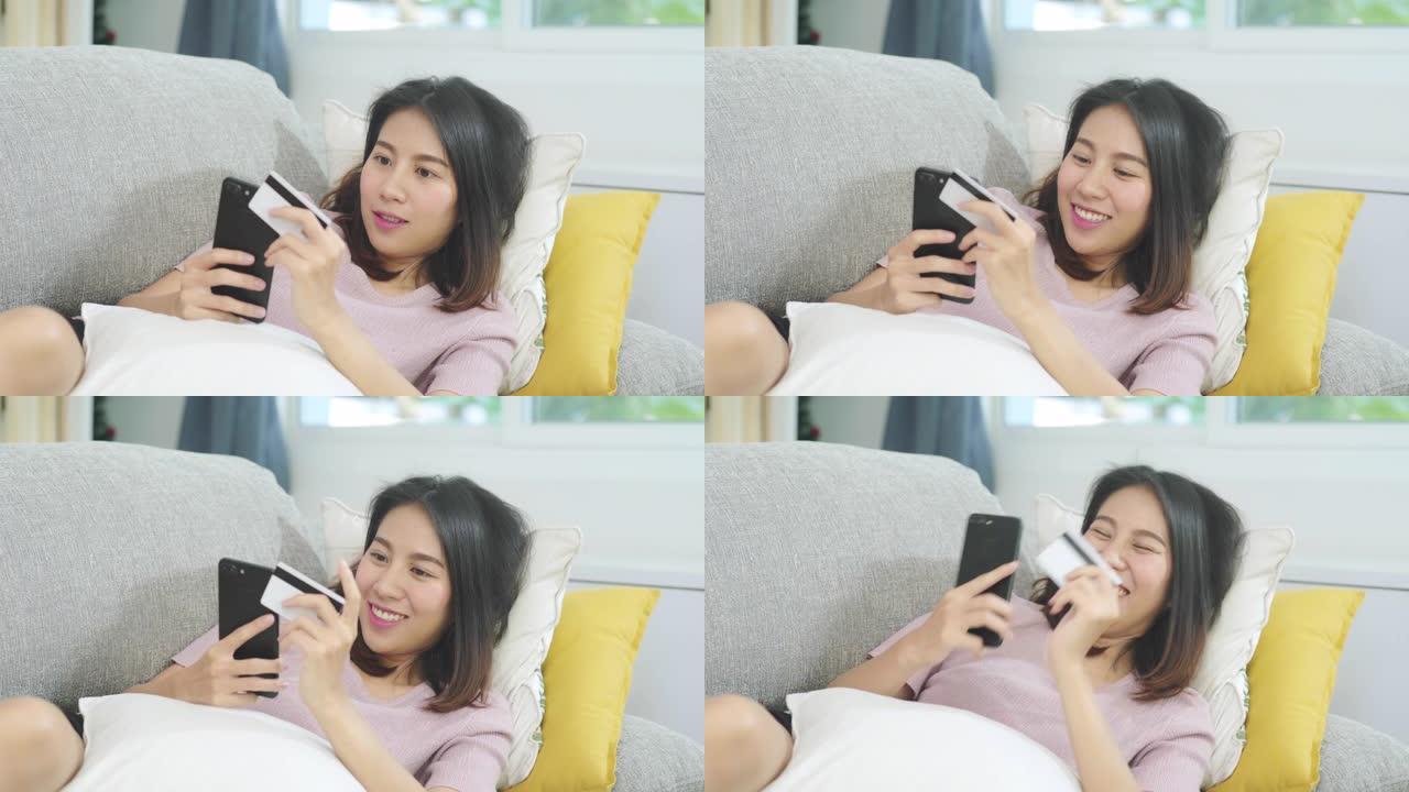 年轻的微笑的亚洲妇女使用智能手机通过信用卡购买网上购物，而躺在沙发上时，在家里的客厅放松。生活方式拉