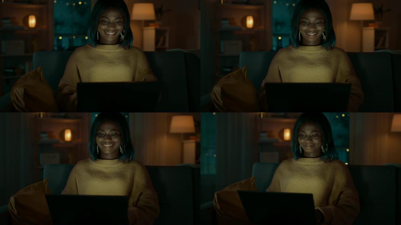 美丽的黑人女孩的肖像坐在家里的沙发上时使用笔记本电脑，她的脸被屏幕灯照亮。可爱的创意女人在镜头前微笑