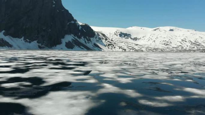 飞越高地湖的水面。南挪威。在部分结冰的湖中可以看到白雪皑皑的岩石和深蓝色的天空。UHD风景优美的空中