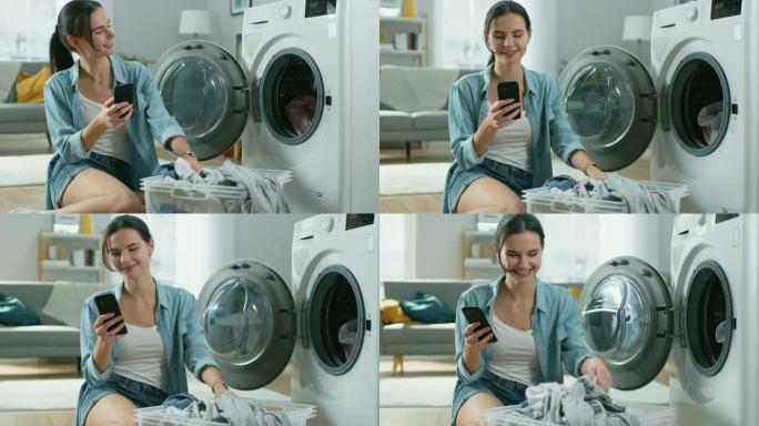 穿着家常衣服的美丽微笑的年轻女子坐在洗衣机前，使用她的智能手机。她给洗衣机装上脏衣服。明亮宽敞的客厅