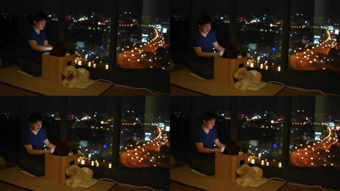 一个亚洲中国人晚上在窗户附近的公寓里玩数字平板电脑在线游戏，可以看到城市景观，玩具狮子狗宠物狗