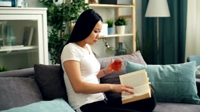 好看的亚洲女人正在看书，享受现代文学，坐在家里的沙发上喝茶放松。教育、千禧一代和房屋概念。