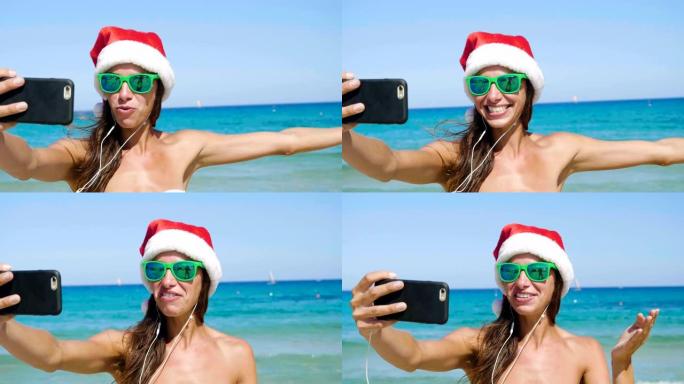 美丽的女孩在手机上自拍，戴着圣诞老人的帽子，戴着墨镜，穿着白色泳衣和白色耳机，背景为海蓝色的水。
