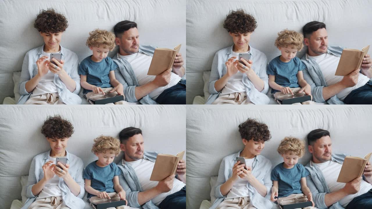 母亲用智能手机儿子拿着平板电脑父亲在家床上看书