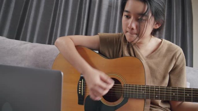 亚洲年轻女孩在家里玩原声吉他和学习在线课程。青少年使用笔记本电脑搜索音乐笔记，在冠状病毒或新型冠状病
