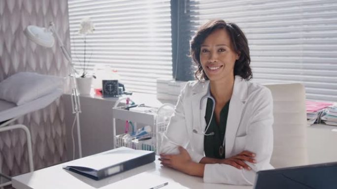 微笑的女医生的肖像穿着白色外套，听诊器坐在办公室的桌子后面