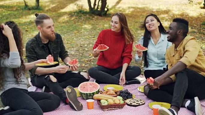 多民族的男人和女人野餐的慢动作吃西瓜，坐在草地上的格子上说话和笑。人与自然的概念。