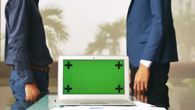 两名商人握手打招呼，并用笔记本电脑绿屏在办公室的窗户附近交谈