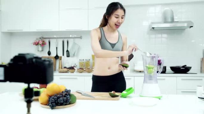 年轻的亚洲女性有影响力的vlogger穿着运动服，在厨房里制作果汁或水果。
