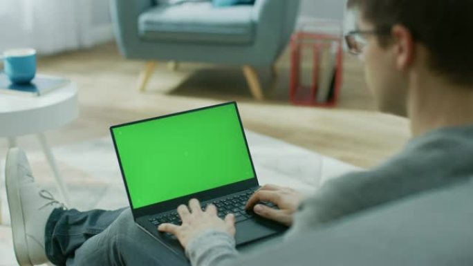 家里的年轻人在带有绿色模拟屏幕的笔记本电脑上工作，在键盘上打字。他坐在舒适客厅的沙发上。肩膀上的镜头
