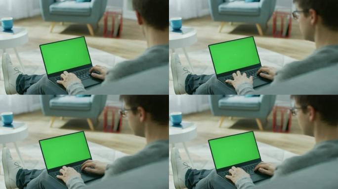 家里的年轻人在带有绿色模拟屏幕的笔记本电脑上工作，在键盘上打字。他坐在舒适客厅的沙发上。肩膀上的镜头