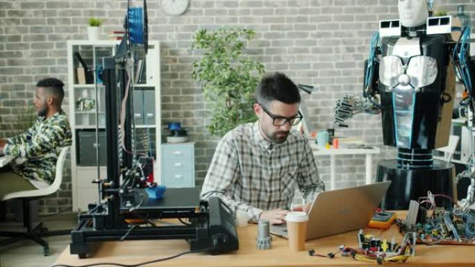 在办公室使用笔记本电脑打字的年轻开发人员，可以看到3d打印机和机器人