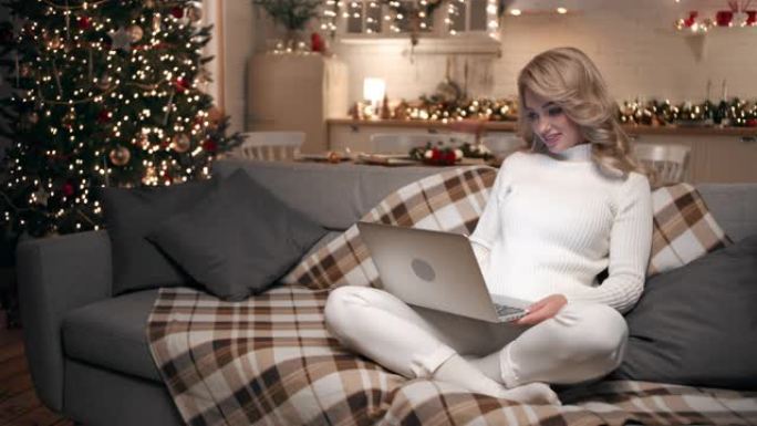 美丽的金发女郎用坐在客厅沙发上的笔记本电脑发送圣诞问候。