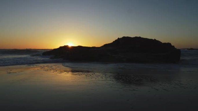 海滩上湿沙的景色和大西洋海浪淹没了它。太阳在葡萄牙日落时滑动一块黑色的岩石。UHD