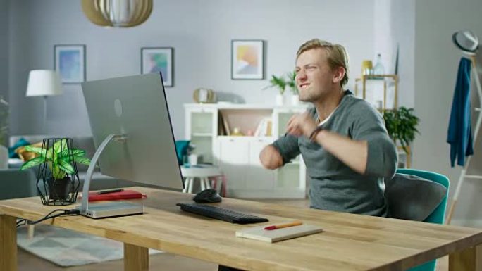 在个人计算机上工作的英俊男子的肖像很幸运，在情感上通过空中打孔来庆祝胜利。背景时尚舒适的客厅。放大镜