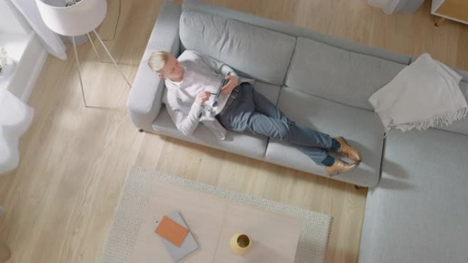 年轻人躺在沙发上，在平板电脑上工作或素描。舒适的客厅配有现代室内、灰色沙发和木地板。顶视图摄像机镜头