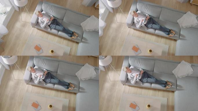 年轻人躺在沙发上，在平板电脑上工作或素描。舒适的客厅配有现代室内、灰色沙发和木地板。顶视图摄像机镜头