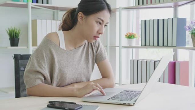 有吸引力的年轻亚洲女性在家工作，看着自由职业者时髦概念的笔记本电脑感到疲倦、无聊和困倦。
