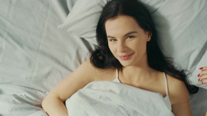 美丽的年轻黑发女人的肖像迷人地躺在床上，性感地微笑着，她的脸被温暖的早晨的阳光照亮。甜美温暖。自上而