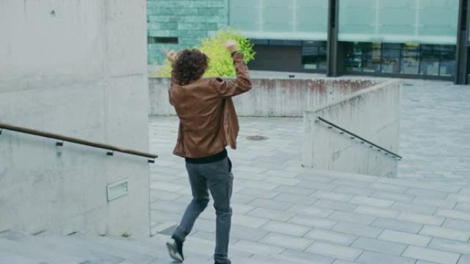 快乐的长发年轻人在走下楼梯时积极跳舞。他穿着一件棕色皮夹克。在商务中心旁边的城市混凝土公园拍摄的场景