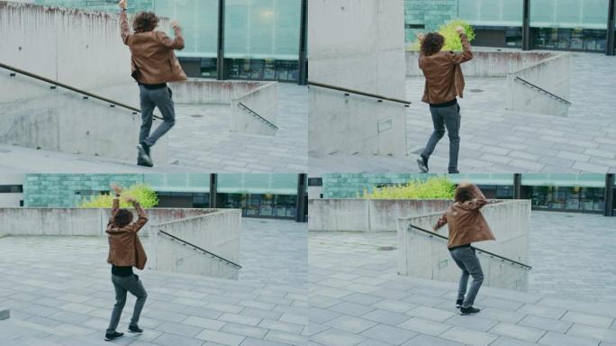 快乐的长发年轻人在走下楼梯时积极跳舞。他穿着一件棕色皮夹克。在商务中心旁边的城市混凝土公园拍摄的场景