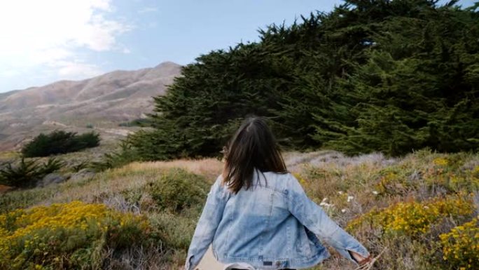 镜头跟随年轻快乐的女人沿着一条小路奔向加利福尼亚大苏尔海岸线的大石头和森林
