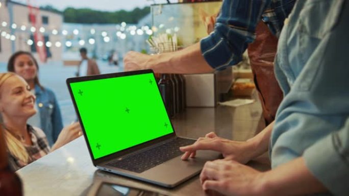 带有绿屏模拟屏幕的笔记本电脑放在出售汉堡和饮料的街头食品卡车或售货亭的柜台上。快乐的顾客收到他们的订